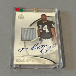 May include: A Michael Huff rookie card in a protective case. The card features a player in a black and white uniform with the number 24. Includes a fabric patch and an autograph. The card is encased in a clear plastic holder.
