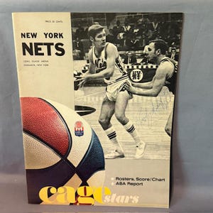 Può includere: Una copertina di un programma di basket vintage dei New York Nets. La copertina presenta un pallone da basket con pannelli rossi, bianchi e blu. Il testo recita "NEW YORK NETS" e "LONG ISLAND ARENA, COMMACK, NEW YORK". Il programma include "Rosters, Score/Chart ABA Report".
