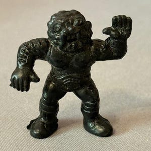 May include: A small, dark-colored figurine of a creature with a humanoid form. The figure has a detailed face, muscular arms, and is wearing boots. The creature is posed with one arm raised.