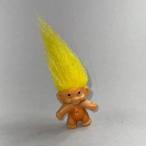 May include: A small, vintage troll doll with a light orange body and a bright yellow, upright hair. The doll has small black eyes and a smiling expression. The troll is standing on a white surface.