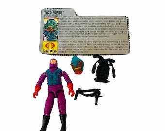 Puede incluir: Una figura de acción de un soldado Cobra Toxo-Viper de la línea de juguetes G.I. Joe. La figura lleva un traje morado con armadura azul y sostiene un arma. La tarjeta dice "Nombre en código: Toxo-Viper, Cobra Hostile Environment Trooper".