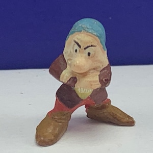 May include: A small plastic figurine of a dwarf with a blue hat, red pants, and a brown jacket. The dwarf has a grumpy expression on his face.