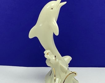 May include: A white porcelain figurine of a dolphin leaping out of the water. The dolphin is painted with gold accents and is standing on a base that resembles waves.