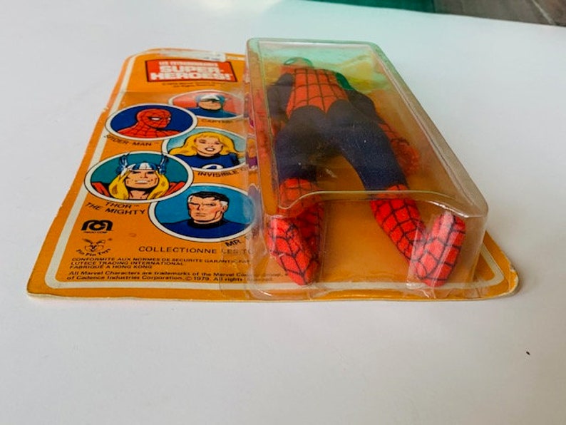 May include: A vintage action figure of Spider-Man in a red and blue suit with a black web design. The figure is packaged in a plastic blister pack with a yellow cardboard backing. The backing features images of other Marvel Super Heroes, including Thor, Invisible Woman, and Spider-Man. The text on the backing reads "Marvel Super Heroes!" and "Collectionne les Super Heros!"