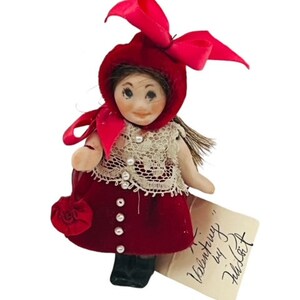 May include: A small doll with a red velvet dress, matching bonnet with a bow, and a lace-trimmed bodice. The doll has a painted face and is holding a small red purse. A tag reads "Valentiny by Felicitat."