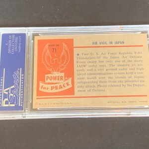 May include: A vintage trading card featuring a photo of two US Air Force Republic F-84 Thunderjets flying over a radar site in Japan. The card has a red, white, and blue design with the words "Power for Peace" and "Air Vigil in Japan".