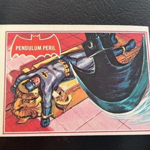 Könnte beinhalten: Eine Vintage-Sammelkarte aus einem Batman-Comic mit einer rot-weiß-blauen Illustration von Batman, der in einer Pendel-Gefahr gefangen ist. Die Karte trägt den Text "Pendulum Peril" in einem weißen Feld mit einem schwarzen Fledermaus-Symbol darüber.