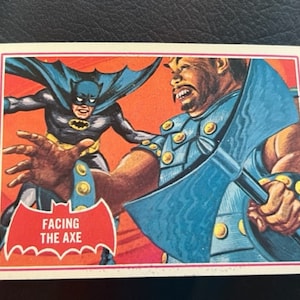 Könnte beinhalten: Eine Comic-Illustration von Batman, der einem großen, blauhäutigen Charakter mit einer Axt gegenübersteht. Der Text "FACING THE AXE" ist auf einem weißen Banner mit einem schwarzen Fledermaussymbol gedruckt.