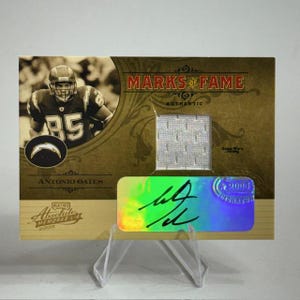 May include: A collectible Antonio Gates football card from the Marks of Fame series. The card features a sepia-toned image of Gates in his Chargers uniform, the text "Marks of Fame", and a jersey swatch.