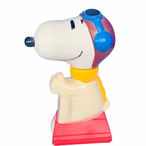 Puede incluir: Una figurita de cerámica de Snoopy vestido de piloto, con un gorro y gafas de aviador marrones y azules, con una bufanda amarilla. Está sentado en una plataforma roja.