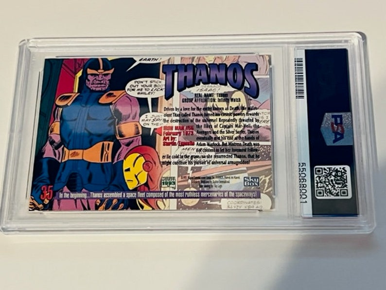 May include: A comic book card featuring Thanos, a purple supervillain from Marvel Comics. The card has a blue and yellow background with the text "THANOS" in large, white letters. The card also includes a small image of Iron Man in the lower right corner.