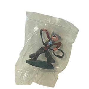 May include: A plastic bag containing a small, painted figurine of a cowboy with a lasso. The cowboy is wearing a brown waistcoat, blue shirt, and brown trousers. The figurine is standing on a green base.