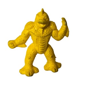 May include: A yellow rubber toy monster with a beak and spiky crest. The monster has a textured surface and is posed with one arm raised.