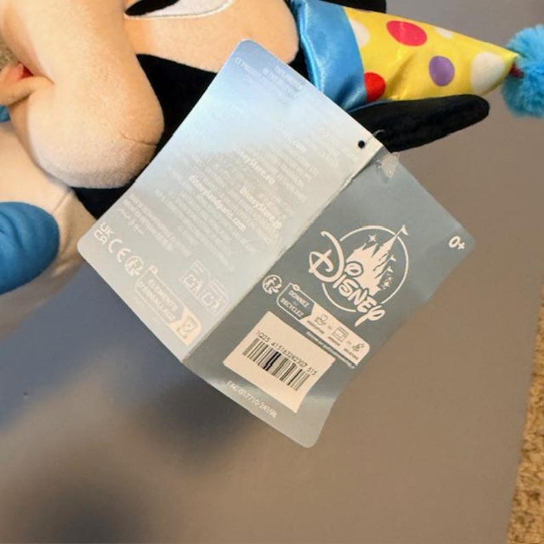 Pode incluir: Um brinquedo de pel&uacute;cia de um personagem da Disney com um chap&eacute;u de festa. O brinquedo tem uma etiqueta com o logotipo da Disney e outras informa&ccedil;&otilde;es. O chap&eacute;u de festa &eacute; amarelo com bolinhas coloridas e um pompom azul.