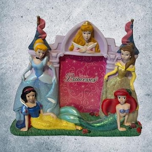 May include: A pink and white castle-shaped picture frame with a Disney Princesses design. The frame features four princesses: Snow White, Cinderella, Aurora, and Ariel. The frame has a place for a photo in the center.