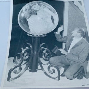 May include: A black and white photograph of a man in a suit kneeling and pointing at a large, round, glass orb on a metal stand. The orb appears to contain a woman's figure.