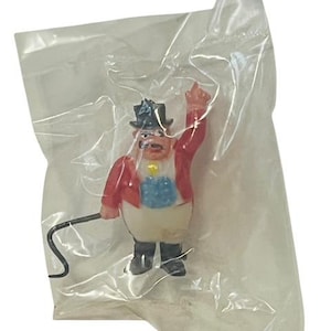 May include: A small figurine of a circus ringmaster in a plastic bag. The ringmaster wears a red jacket, black top hat, and white trousers. He is holding a whip and has his arm raised.