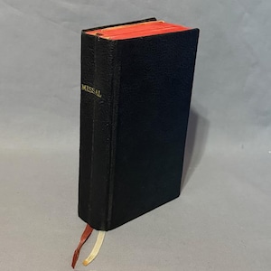 May include: A black book with the word "MISSAL" in gold lettering on the spine. The edges of the pages are red, and two ribbon bookmarks are visible. The book is standing upright against a gray background.
