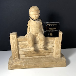 Puede incluir: Una figurita de cerámica beige de un niño con gorra sentado en una valla. La figurita está hecha por Royal Haeger, una empresa que lleva en funcionamiento desde 1871.
