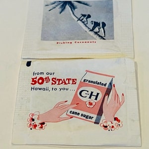 Puede incluir: Dos paquetes de papel blanco con una ilustración roja y azul de manos sosteniendo una caja de azúcar de caña granulada. La caja tiene el texto "from our 50th STATE Hawaii, to you... CH cane sugar" impreso en ella. El paquete superior tiene una ilustración en blanco y negro de dos personas recogiendo cocos de una palmera con el texto "Picking Coconuts" impreso debajo.