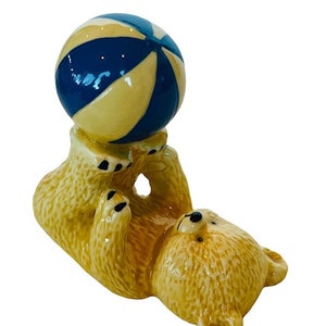 Può includere: Una statuetta in ceramica di un orso giallo che bilancia una palla da spiaggia blu e gialla sulle sue zampe.