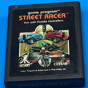 May include: A black Atari game cartridge with a colorful illustration of cars and a motorcycle on the front. The game title is "STREET RACER" and the text "Use with Paddle Controllers" is below the title. The text "game program" is at the top of the cartridge. The text "ATARI" and "CX2612" are at the bottom of the cartridge. The text "Label, Program & Audiovisual © 1978 ATARI, INC." is below the text "CX2612".