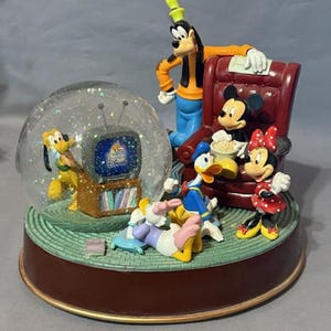 May include: A Disney snow globe featuring Goofy, Mickey Mouse, Minnie Mouse, Donald Duck, Daisy Duck, and Pluto. The characters are gathered around a TV, with a red armchair and popcorn. The base is brown with a green mat.
