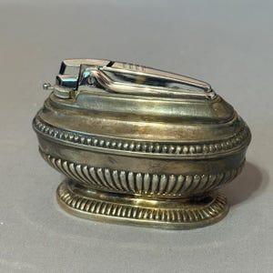 May include: An antique silver-toned lighter with a rounded body and a hinged top. The lighter has a decorative design with a series of small, raised dots and vertical lines. The base is also silver-toned and has a similar decorative pattern.