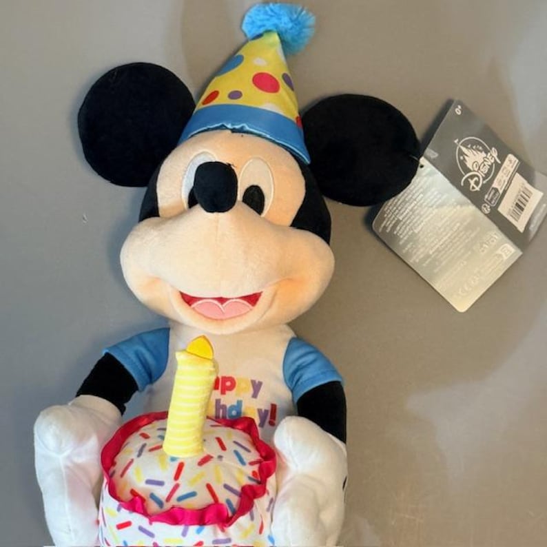 Pode incluir: Um brinquedo de pel&uacute;cia do Mickey Mouse usando um chap&eacute;u de festa e segurando um bolo de anivers&aacute;rio. O bolo &eacute; branco com granulado colorido e uma vela amarela. A camisa diz "Happy Birthday!" em letras amarelas. O brinquedo tem orelhas pretas e uma etiqueta da Disney.