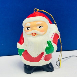May include: A vintage Christmas ornament featuring a Santa Claus figure with a white beard and red suit. He is holding a bag of gifts and wearing a green hat. The ornament is made of ceramic and has a gold string for hanging.