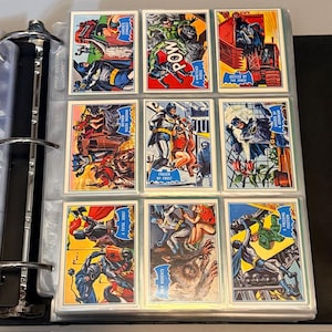 Könnte beinhalten: Eine Sammlung von Batman-Sammelkarten im Vintage-Stil in einem Ordner. Die Karten zeigen farbenfrohe Comic-Illustrationen von Batman in verschiedenen Actionposen, mit Sprechblasen und Bildunterschriften. Der Ordner ist schwarz mit einem Metallring.