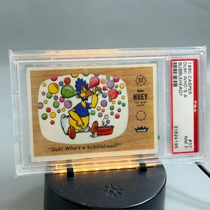 May include: A 1960 Fleer Baby Huey trading card encased in a PSA slab. The card features a cartoon duck blowing colorful bubbles with the text "Duh! Who's a bubblehead?" and "37 Baby HUEY THE BABY GIANT". The slab label includes the PSA logo and serial number.