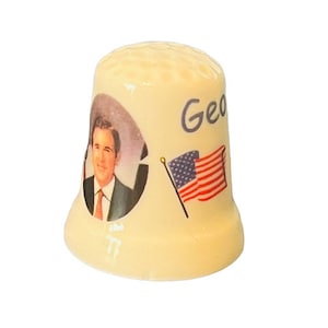 May include: A white ceramic thimble with a picture of George W. Bush and an American flag. The thimble has the word "Geo" written on it.