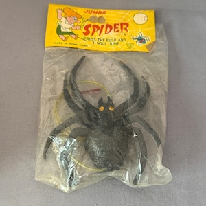 May include: A black plastic toy spider in a clear plastic package with a yellow header. The header reads "JUMBO SPIDER" with the text "PRESS THE BULB AND I WILL JUMP". The spider has yellow eyes.