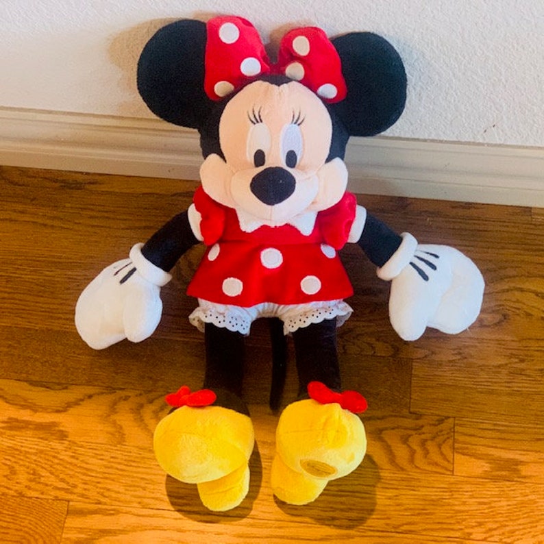 May include: A plush Minnie Mouse toy with a red polka dot dress, white gloves, and yellow shoes. The toy has a black nose and large black eyes.