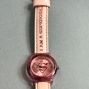 May include: Pink Supergirl watch with a light pink leather band. The watch face features the Supergirl logo in red and white. The band is printed with the words "I AM A SUPERGIRL".