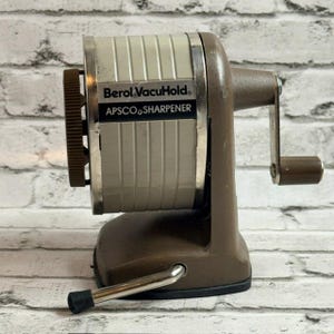 May include: A vintage Berol VacuHold APSCO pencil sharpener featuring a beige cylindrical body with a brown base and handle. The front label displays the text Berol VacuHold and APSCO SHARPENER. It includes a manual crank and a metal lever.