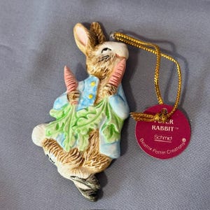 Puede incluir: Un adorno de cerámica de Peter Rabbit con una chaqueta azul, sosteniendo zanahorias y verduras de hoja verde. El adorno tiene una cuerda dorada para colgar y una etiqueta roja que dice "Peter Rabbit" y "Beatrix Potter Creations".
