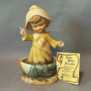 May include: A porcelain figurine of a child in a boat, wearing a yellow outfit and hat. The figurine has a white and tan sash and is pointing with one hand. A tag reads "Little Bible Friends".