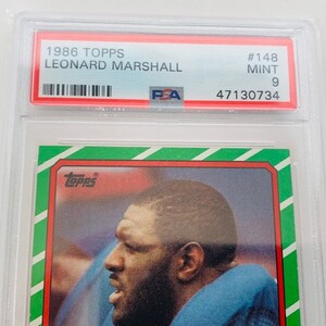 Op de afbeelding: Een Topps-verzamelkaart uit 1986 met Leonard Marshall, een American footballspeler. De kaart is in mint conditie en heeft een PSA-beoordeling van 9. De kaart is groen, rood en wit met het Topps-logo in de linkerbovenhoek.