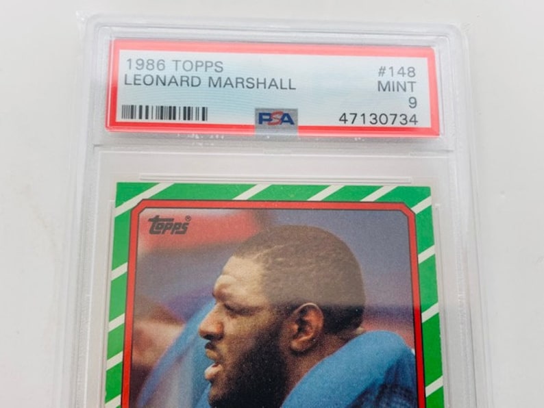 Op de afbeelding: Een Topps-verzamelkaart uit 1986 met Leonard Marshall, een American footballspeler. De kaart is in mint conditie en heeft een PSA-beoordeling van 9. De kaart is groen, rood en wit met het Topps-logo in de linkerbovenhoek.