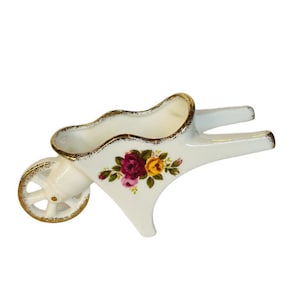 May include: White ceramic wheelbarrow with a gold rim and a floral design featuring pink, red, and yellow roses.