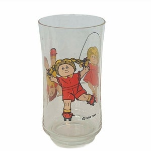 May include: A clear glass drinking glass with a colorful image of three Cabbage Patch Kids dolls wearing red dresses and roller skates. The text "©1984  O.A.A." is printed on the bottom of the glass.