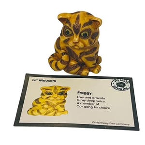 May include: A small ceramic figurine of a yellow and brown tabby cat with black stripes. The cat is sitting with its tail wrapped around its legs. The figurine is part of a collection called "Lil' Mousers" and is named "Froggy". The card reads "Low and gravelly is my deep voice. A member of our gang by choice."  © Harmony Ball Company