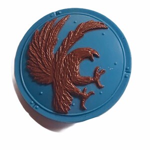 May include: A round blue shield with a brown eagle design. The eagle is depicted with its wings spread and talons extended.