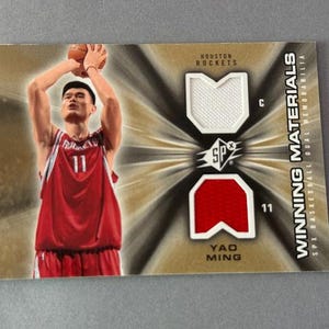 Puede incluir: Una tarjeta de baloncesto con Yao Ming con la camiseta roja de los Houston Rockets, número 11. La tarjeta incluye muestras de camiseta y el texto "Winning Materials".