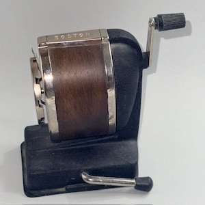 May include: A vintage black and brown pencil sharpener with the word "BOSTON" on the top. The sharpener has a silver metal trim and a crank handle. The base is rectangular with a silver metal pencil rest.
