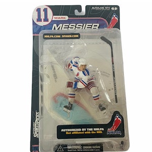 May include: A Mark Messier action figure in a clear plastic package. The figure is wearing a white, blue, and red hockey uniform. The packaging includes the text "11 MARK MESSIER" and "SPORTSPICKS". The figure is authorised by the NHLPA.