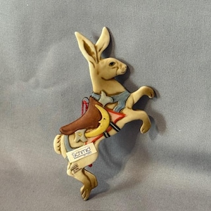 Puede incluir: Un adorno de cerámica blanca de conejo con una silla de montar azul y roja y una luna creciente amarilla. El adorno está hecho por Schmid y tiene una cuerda roja para colgar.