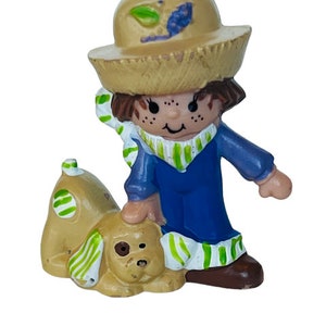 May include: A small plastic figurine of a girl wearing a straw hat, blue overalls, and a green and white striped scarf. She is holding a small brown dog with green and white spots.